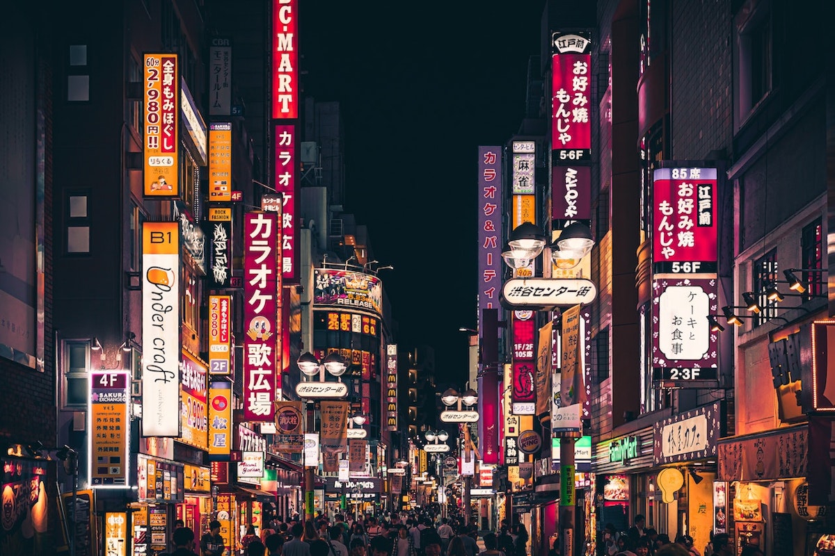 A vibrant, illuminated city street at night is filled with colorful neon signs and billboards in Japanese. The street is bustling with people, and the atmosphere is energetic. Various shops and eateries line the sides of the street, extending into the distance.