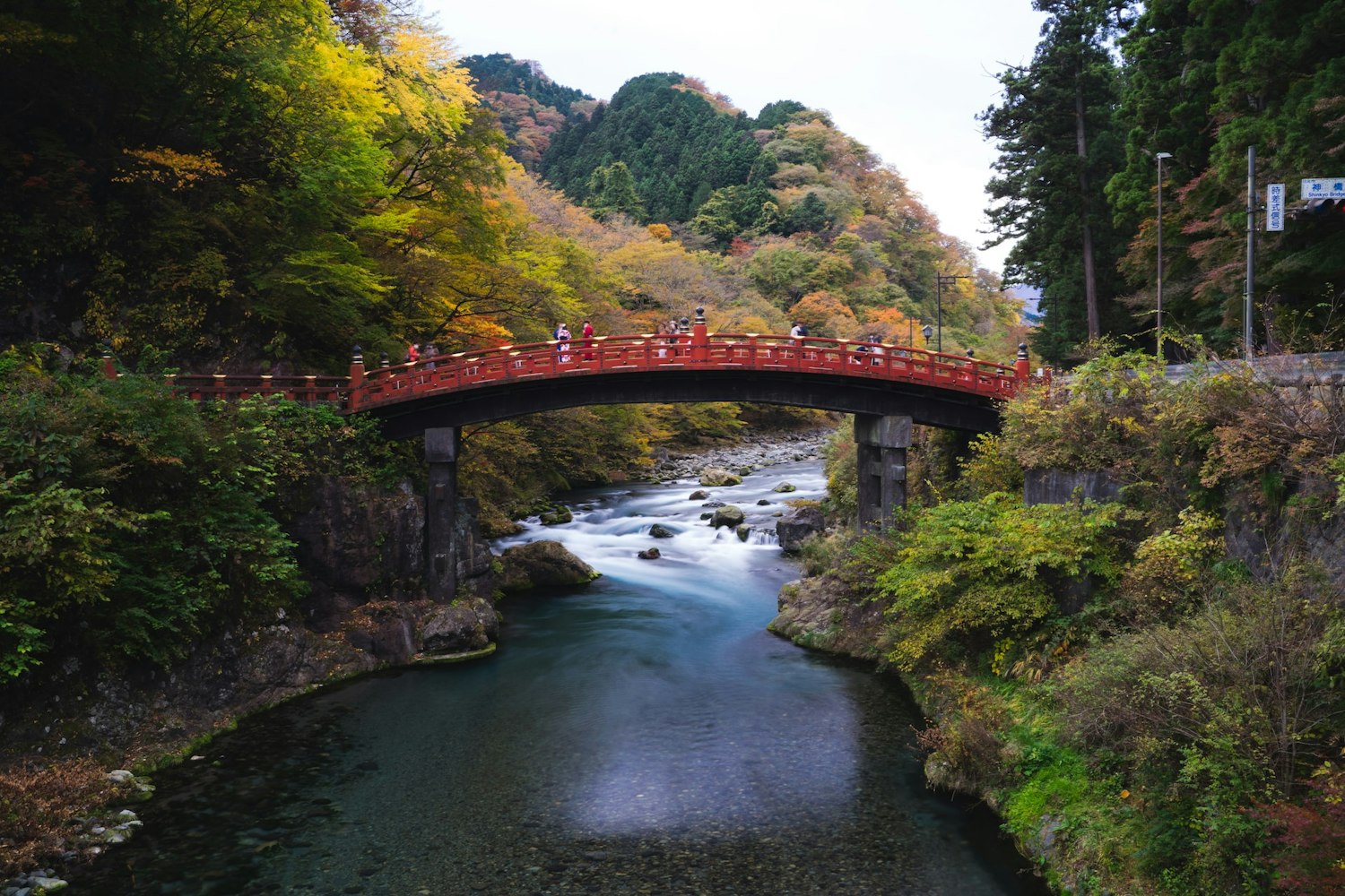 A picturesque red bridge arches over a tranquil river surrounded by lush, multicolored autumn foliage in a forested area. People are seen walking across the bridge, and hills covered in dense trees form a scenic backdrop. The sky is overcast.