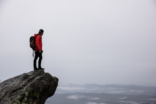A person in a red jacket, black hat, and backpack stands on the edge of a cliff overlooking a vast, cloudy landscape. Fog covers the distant mountains, creating a moody and serene atmosphere. The sky is overcast, adding to the dramatic scene.
