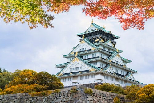 A traditional Japanese castle with white walls and teal roofs sits atop a stone base, surrounded by vibrant foliage in shades of green, yellow, and red. The sky above is mostly cloudy with hints of blue peeking through.