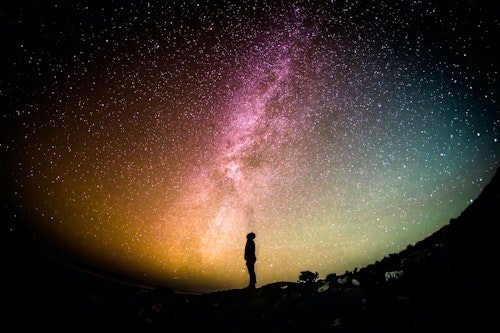 Silhouetted person stands on a hill, looking up at a vibrant, star-filled night sky. The Milky Way is prominently visible, creating a colorful, luminous pathway across the sky. The horizon glows with a gradient of warm and cool tones.