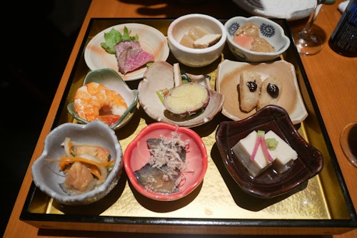 A tray of assorted Japanese appetizers, each in its own small dish. The selection includes seafood, vegetables, tofu, and pickled items, all artfully arranged. The dishes vary in colors and shapes, contributing to a visually appealing presentation.
