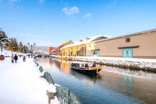 A boat with people is floating along a snow-covered canal, flanked by snowy sidewalks and historic brick buildings under a clear blue sky. A few people are walking on the left path, and trees are visible in the distance.