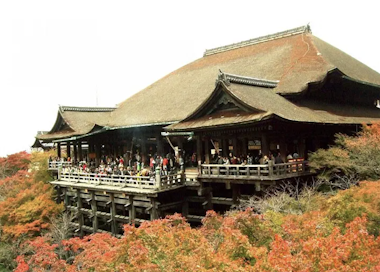 The image shows Kiyomizu-dera, a historic Buddhist temple in Kyoto, Japan. It features a large wooden stage supported by pillars and surrounded by vibrant autumn foliage. Many visitors are gathered on the terrace.